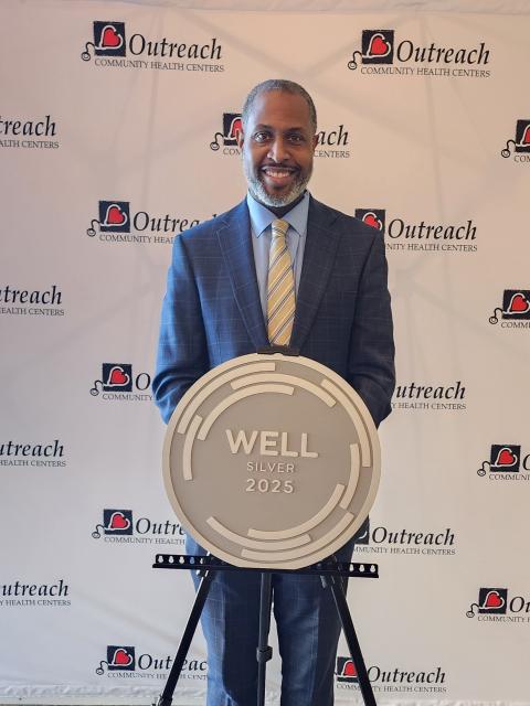 Person in a blue suit stands behind a large circular “WELL Silver 2025” certification plaque displayed on an easel, in front of a backdrop featuring Outreach Community Health Centers logos.