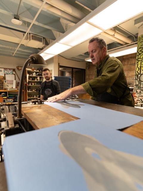 An artist operates a letterpress printing machine while another person observes in a printmaking studio.