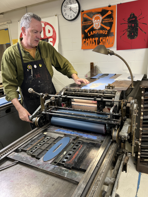 An artist adjusts paper on a vintage letterpress printing press in a studio displaying colorful poster prints on the wall.