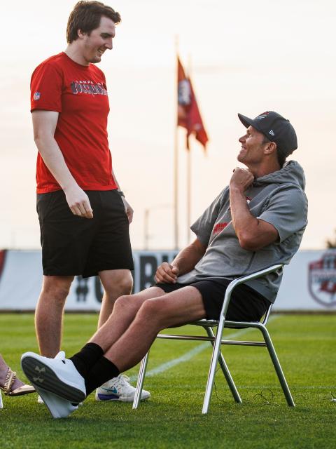 A person wearing a Tampa Bay Buccaneers T-shirt stands on a practice field talking with a seated football player during a team event.